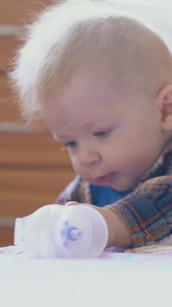 funny little boy in warm shirt nibbles part of inhaler leaning on large soft bed in light children room close view