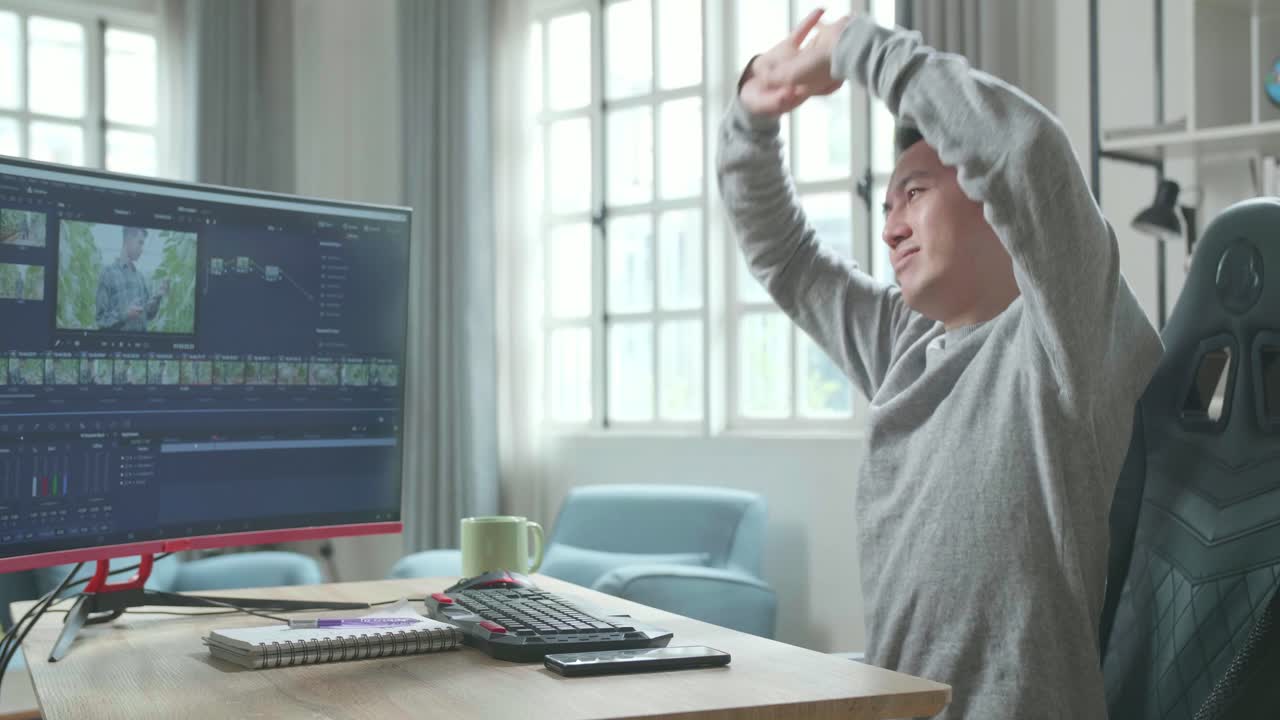 Asian Video Editor Man In Long Sleeved T-Shirt Stretching And Leaning Back While Using Desktop Computer For Editing Video At Home.