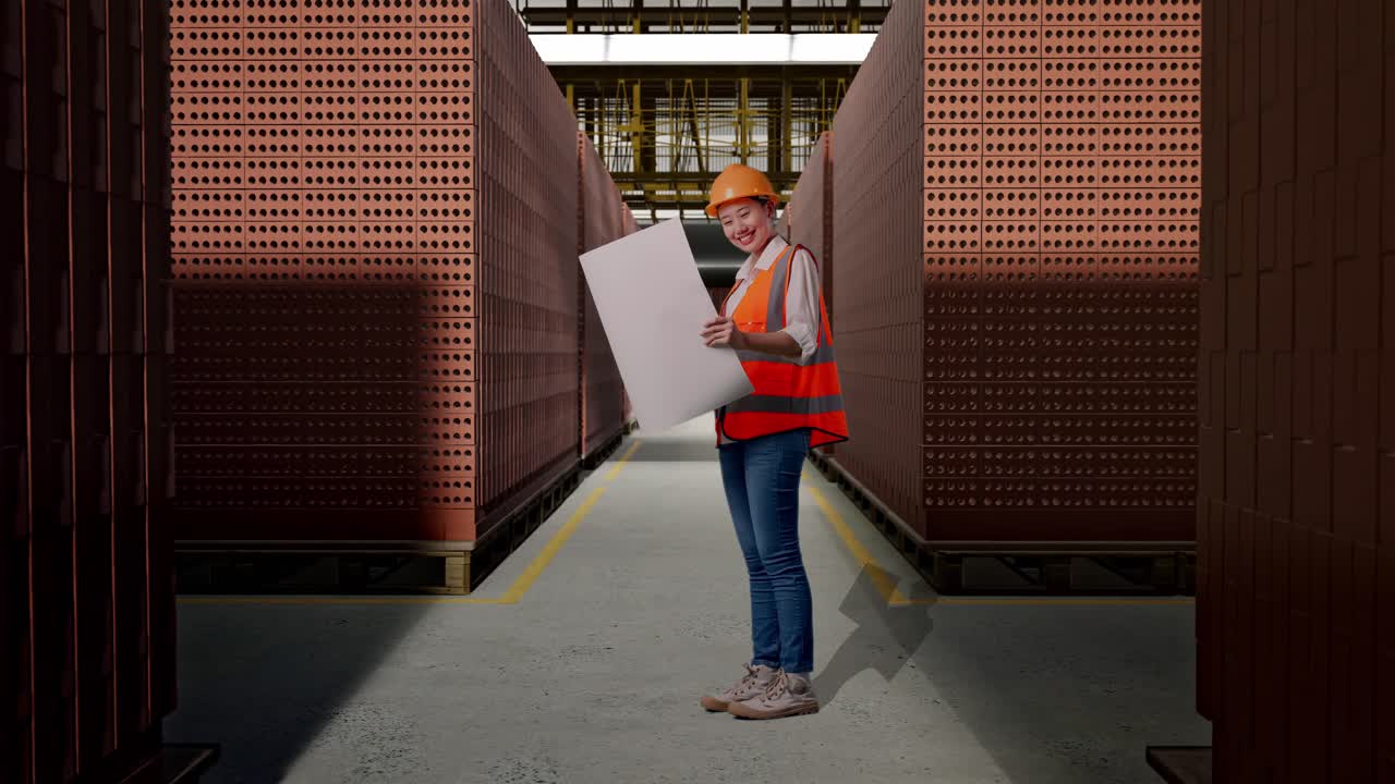 Full Body Side View Of Asian Female Engineer With Safety Helmet Looking At Blueprint In Her Hands While Standing With Red Brick Packed in Stacks Are Stored