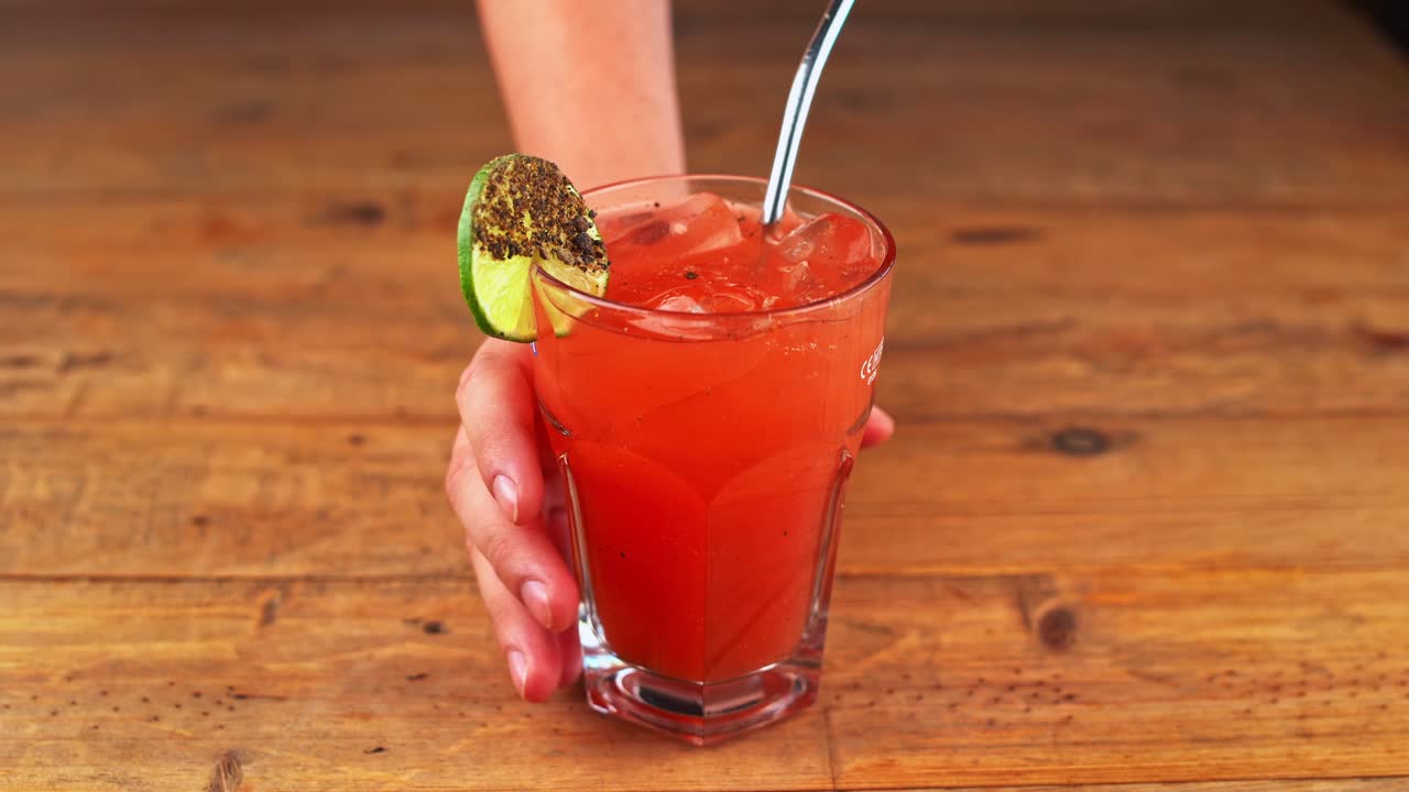 pessoa removendo da mesa de madeira um coquetel bloody mary, com limão e uma palha