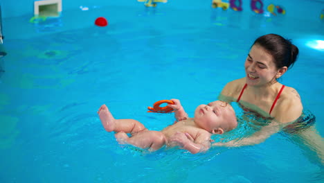 smiling woman supporting a baby lying on the water surface in the swimming pool. female coach having a lesson of swimming with a kid.