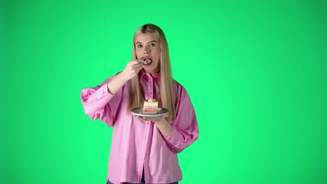 pretty young blonde woman enjoying eating delicious cake dessert, studio portrait with green background