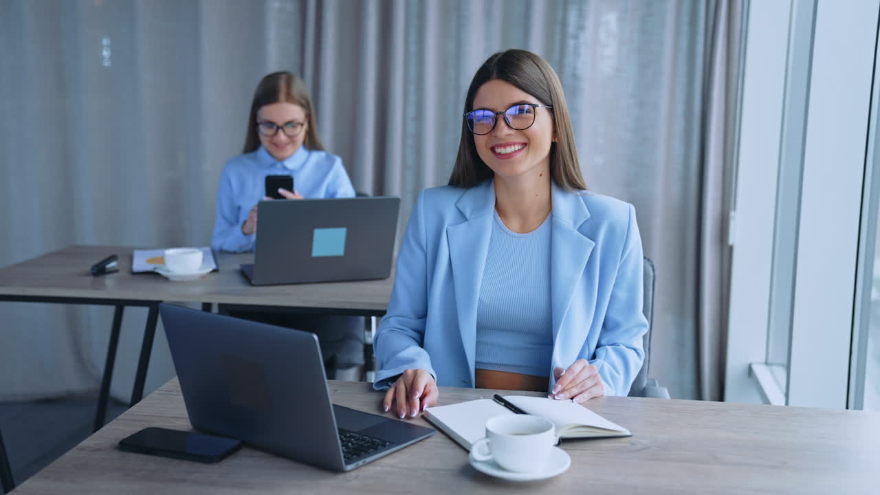 Happy female office employees working in a light room. Women sit at desks using laptops and smartphones. Blurred backdrop.