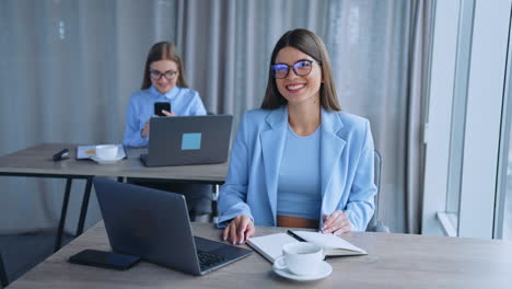 happy female office employees working in a light room. women sit at desks using laptops and smartphones. blurred backdrop.