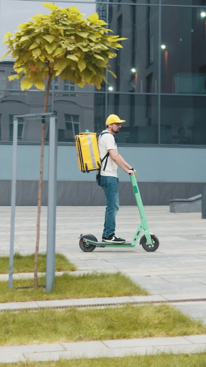 A delivery person rides an electric scooter through a city carrying a yellow insulated bag