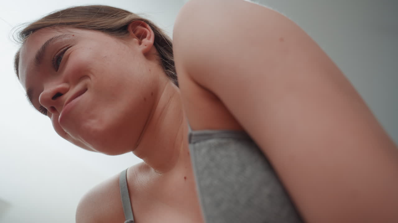 Donna incinta latina che respira nella stanza, ritratto ravvicinato dal basso che mostra una canotta grigia, sudore sulla pelle, sorriso sottile ed espressione determinata, atmosfera intima che trasmette attesa.