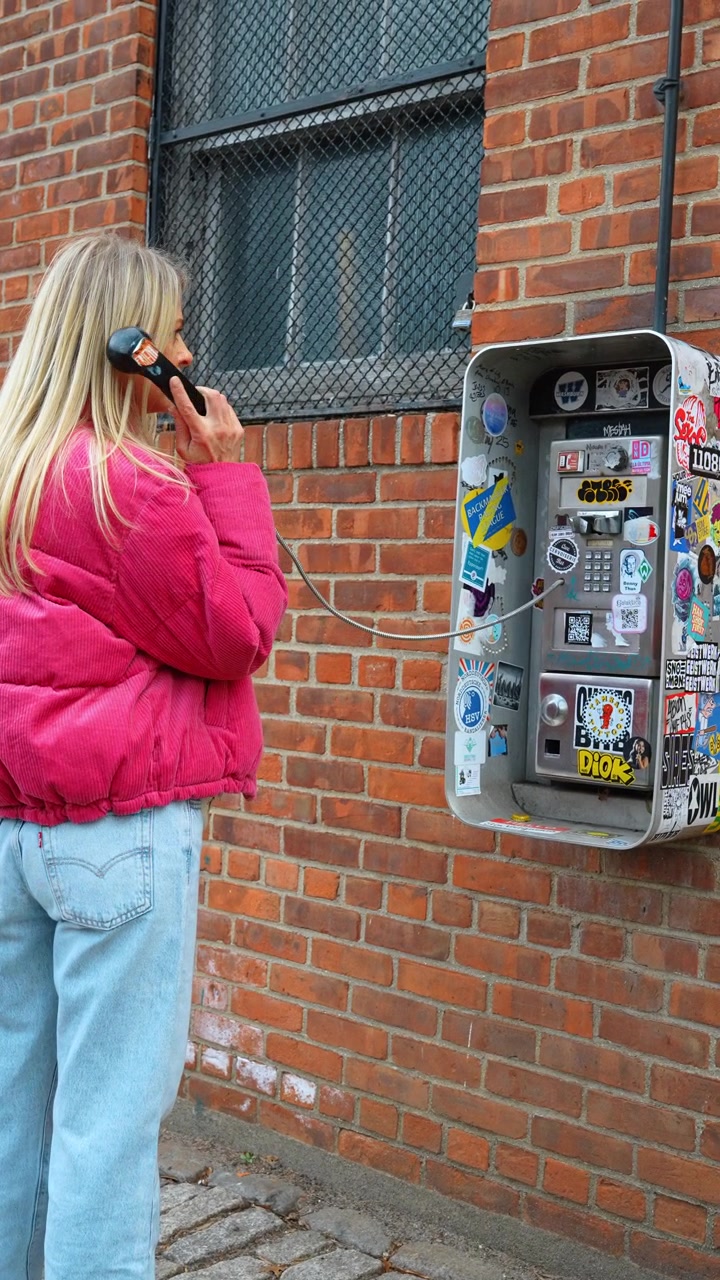 New York, USA, 25 maggio 2026: Donna che usa un telefono pubblico vintage su un muro di mattoni rossi a New York. Vista di profilo di una donna bionda con una giacca rosa che tiene la cornetta del telefono presso una cabina telefonica coperta di adesivi.