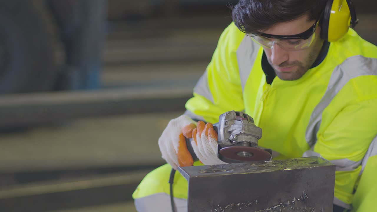 trabajador masculino caucásico en un uniforme seguro usar soldadores cueros, rueda eléctrica molienda en la estructura de acero en el factor en el garaje, chispas bokeh naranja vuelan a los lados