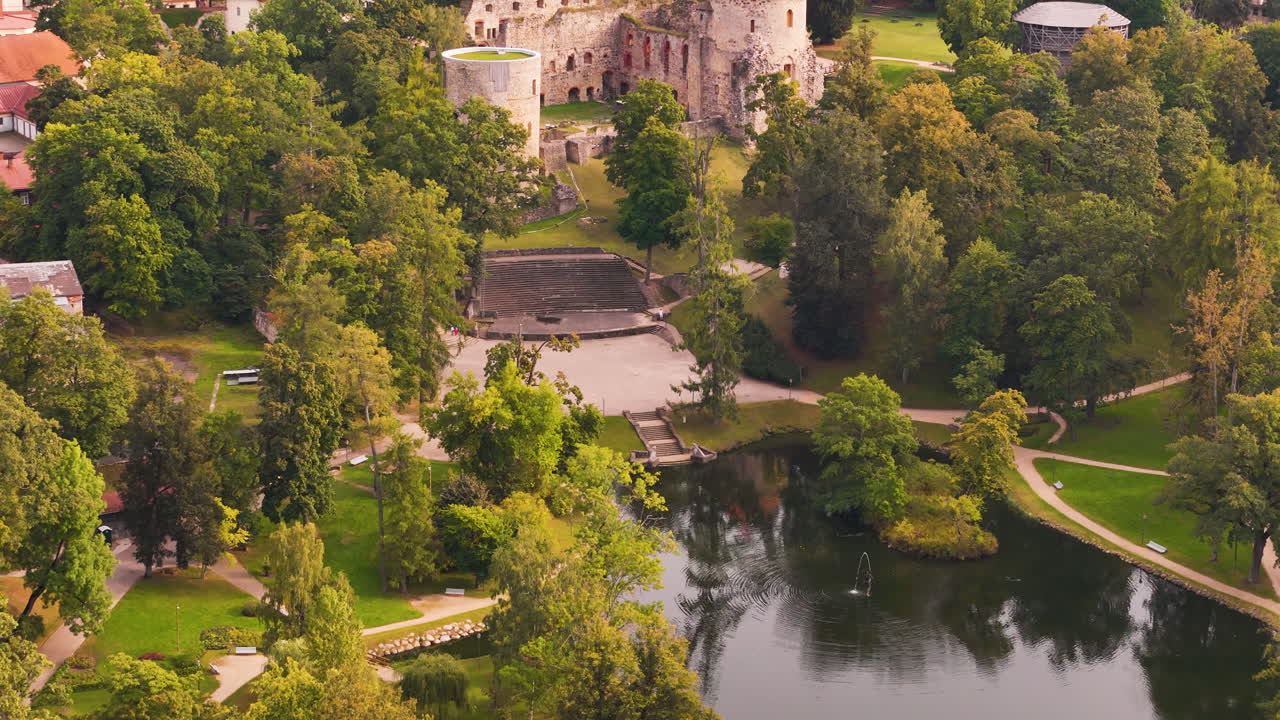 Drone footage of Cesis Medieval Castle ruins, lush greenery, and a tranquil pond in Latvia.