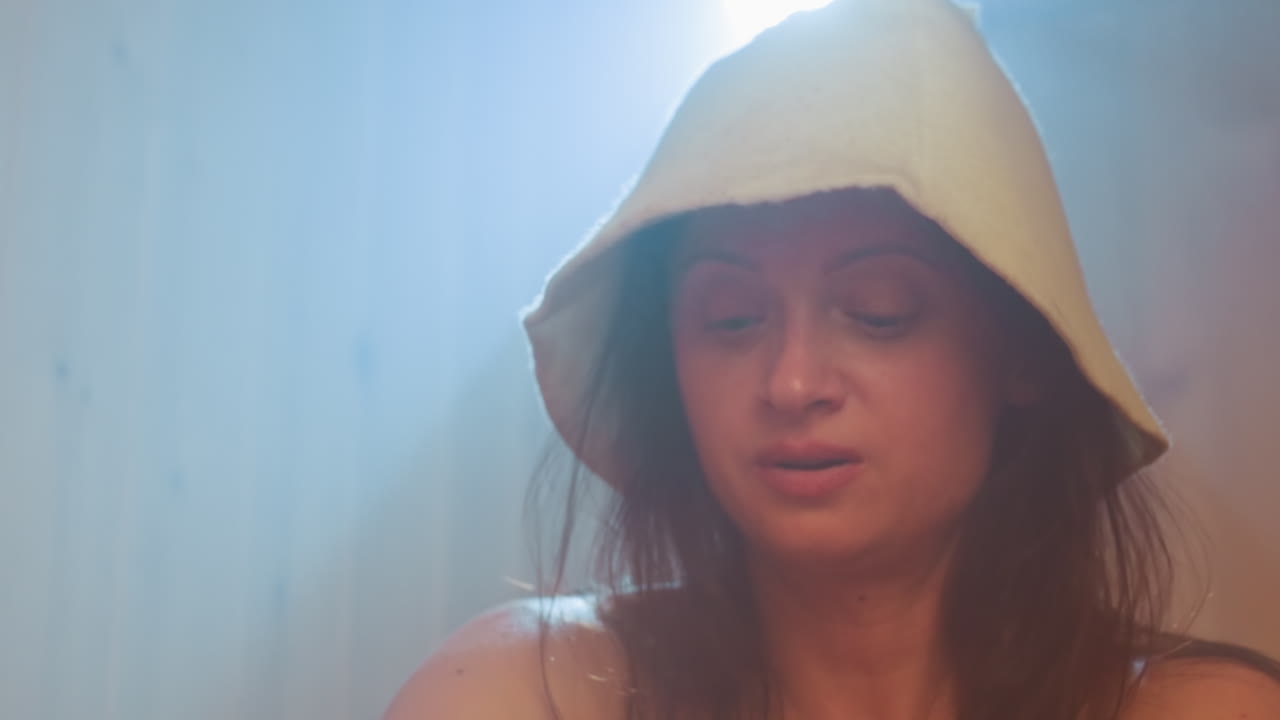 misty spa scene with young woman wearing felt spa hat illuminated by soft backlight gently massaging cream into leg during serene self care ritual in wooden steam cabin ambience