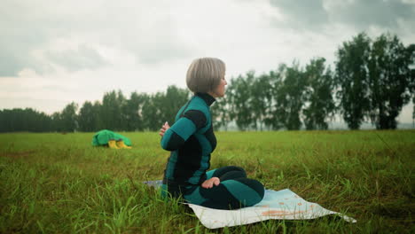 side view of middle-aged woman sitting on mat in sukhasana pose, practicing yoga with eyes closed, under a cloudy sky with trees lined up and a yellow boot and cloth in the background