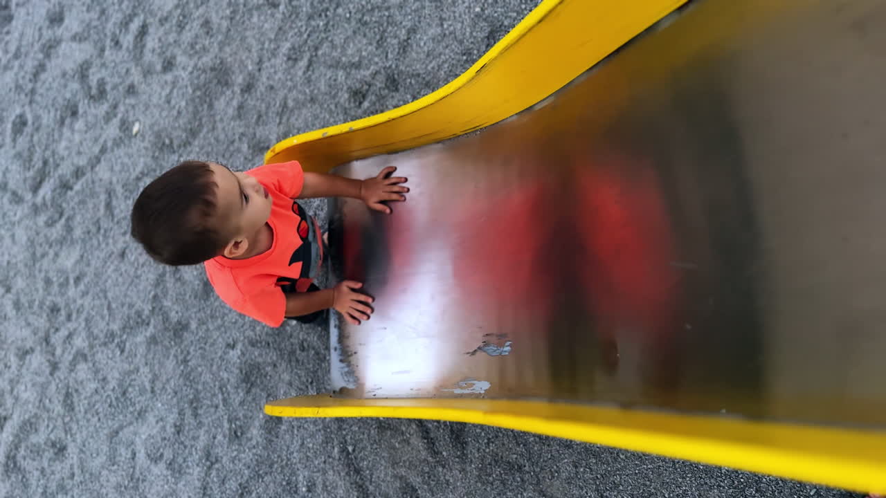 Dark-haired toddler boy tries to climb up by the metal slide. Happy kid jumps up and walks away. Top view. Vertical screen.