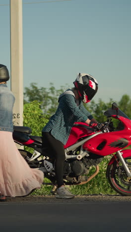 dos mujeres con cascos, una sentada en una bicicleta deportiva roja mientras la otra se baja de la bicicleta, están en la carretera, con coches pasando en el fondo
