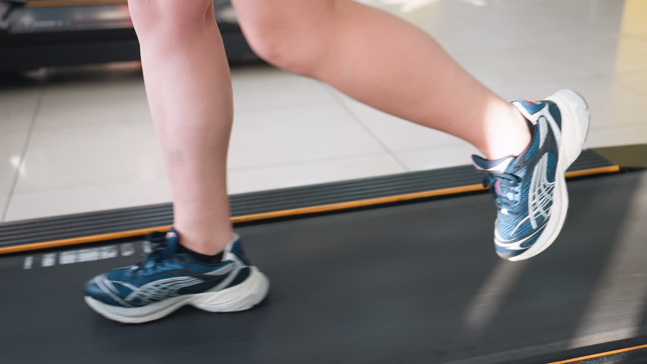 side view of person legs running on treadmill wearing blue sneakers during indoor workout session on tiled gym floor with additional fitness equipment and impulse machine visible in background