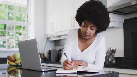 Eine-Afroamerikanische-Frau-Macht-Sich-Notizen-Und-Benutzt-Einen-Laptop-In-Der-Küche,-Während-Sie-Von-Zu-Hause-Aus-Arbeitet