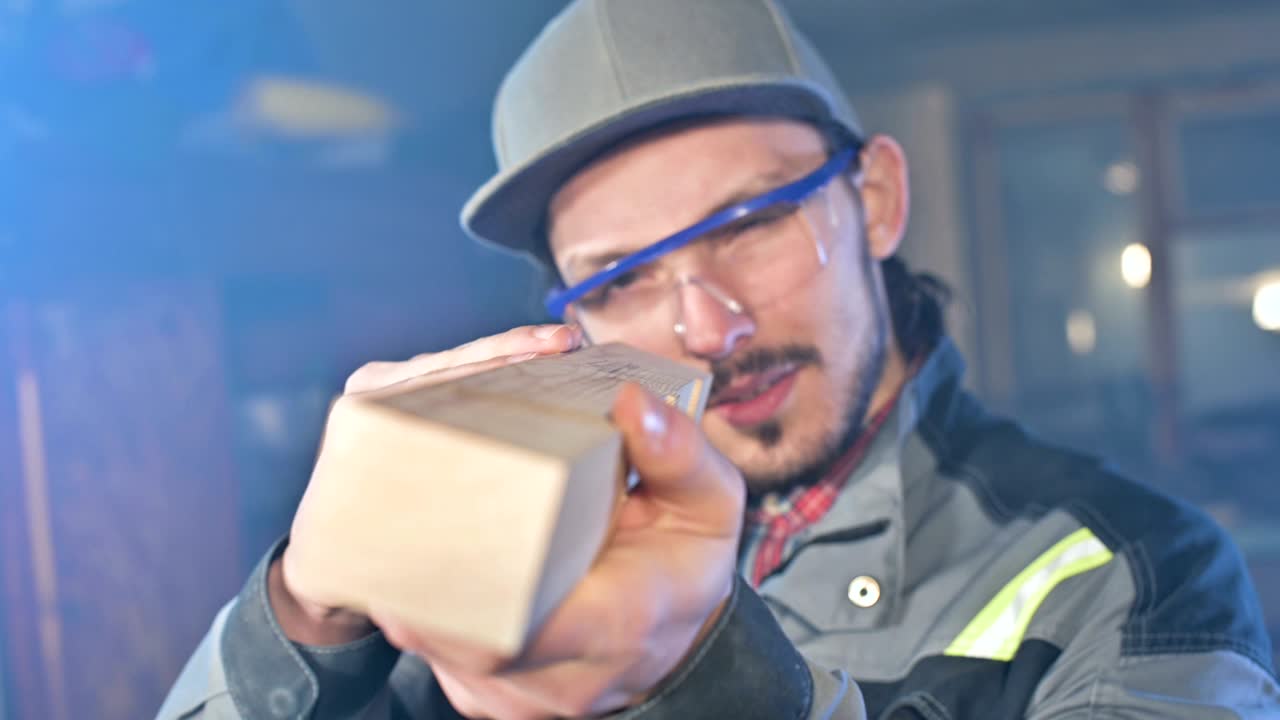 joven carpintero barbudo concentrado con gafas de protección y una gorra comprueba la pieza de trabajo
