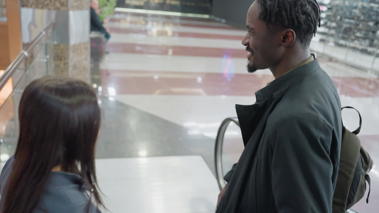 Black man interacting with white woman as they step off escalator into open hallway of contemporary shopping mall, engaging in calm conversation under bright indoor lights