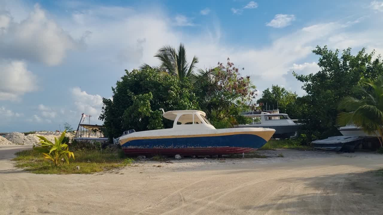 Barca blu e gialla a terra a Maafushi, Maldive, probabilmente per manutenzione o stoccaggio, circondata da vegetazione tropicale in una giornata ventosa, catturando la vita isolana e l'atmosfera costiera