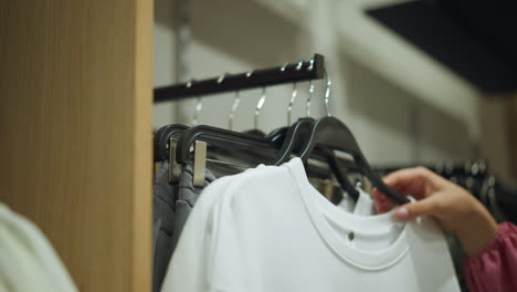 una mano en tela rosa devuelve cuidadosamente una tapa blanca a un estante de ropa con pantalones de ceniza, el fondo muestra una vista borrosa de ropa bien arreglada en los estantes con un entorno de tienda bien iluminado