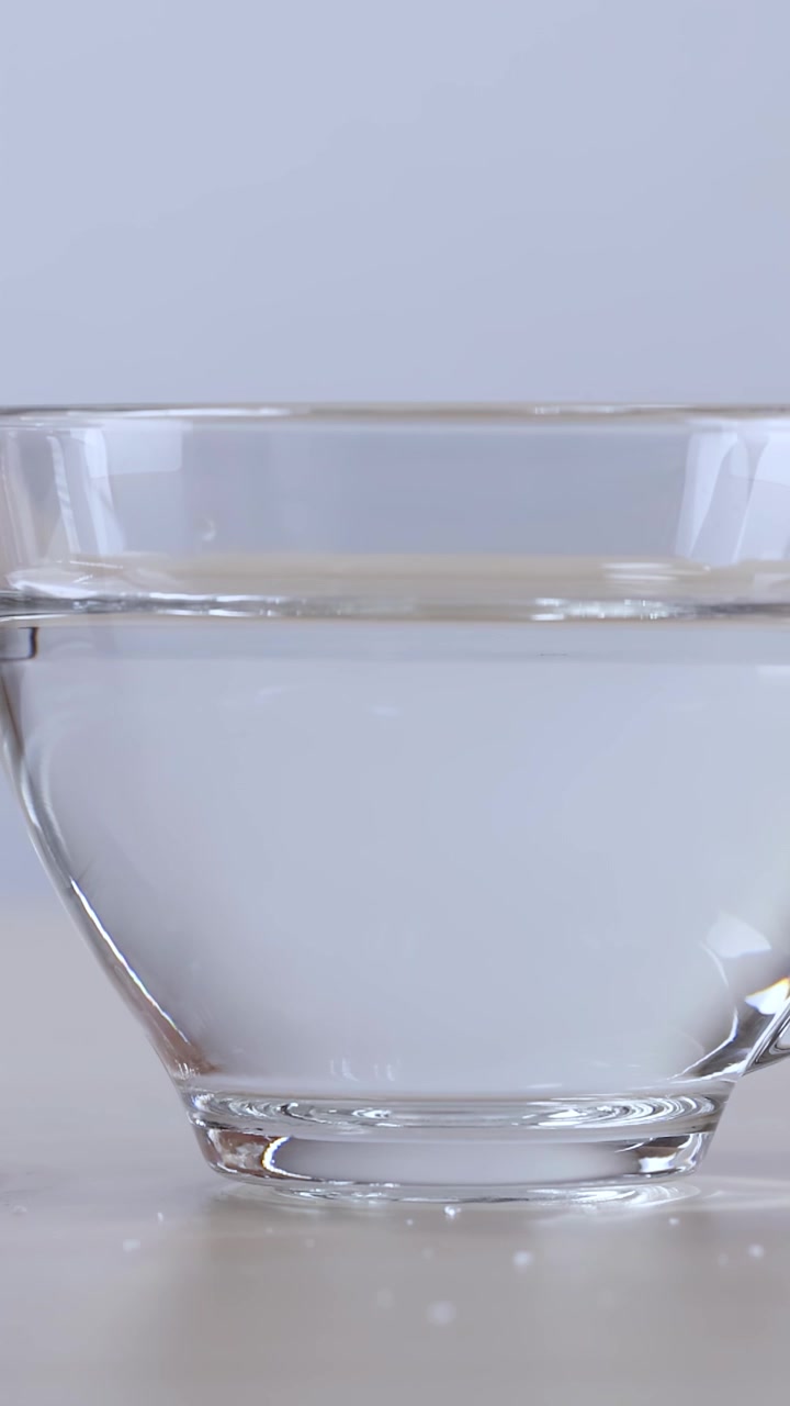 Una tazza di vetro trasparente piena d'acqua scivola nell'inquadratura su una superficie di legno.