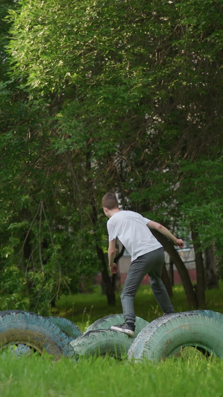 vista posterior en cámara lenta de un niño en una camisa blanca y pantalones grises mientras salta a través de neumáticos dispuestos en un campo de hierba, rodeado de árboles exuberantes en el fondo