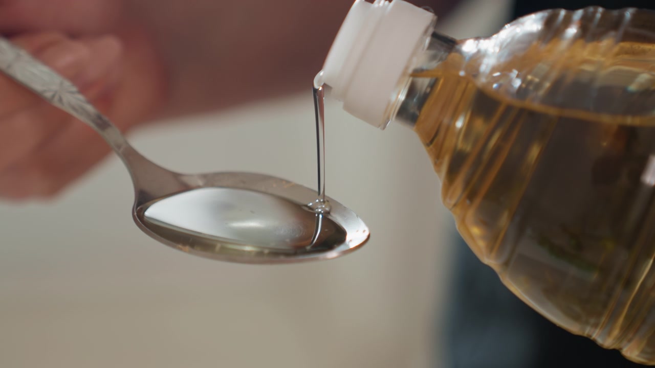 Una mano inclina una piccola bottiglia di plastica per versare liquido ambrato in un cucchiaio di metallo, mostrando un flusso visibile, la formazione di una goccia, superfici riflettenti e un movimento di dosaggio attento sotto una luce interna soffusa con messa a fuoco superficiale.