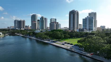 driving car on road at river in front of american skyline. sunset time in st. petersburg, florida. residential tower and skyscraper of downtown. palm trees in park. aerial backwards wide shot.