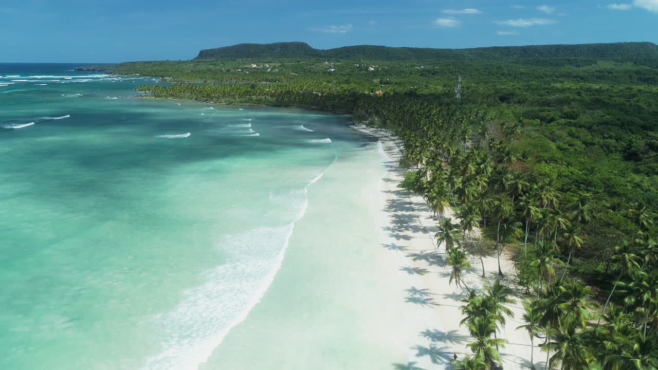 Drone che scende dritto verso una spiaggia con palme, onde e acqua cristallina