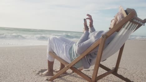 Mujer-Mayor-Hispana-Relajándose-En-Una-Tumbona-En-La-Playa-Al-Atardecer,-Usando-Un-Teléfono-Inteligente