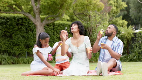 Happy-african-american-parents,-daughter-and-son-blowing-bubbles-sitting-in-garden,-slow-motion