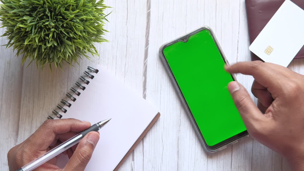 Person working on a table with a green screen phone, notebook, pen, and credit card