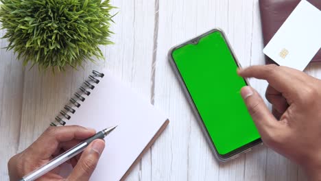 person working on a table with a green screen phone, notebook, pen, and credit card