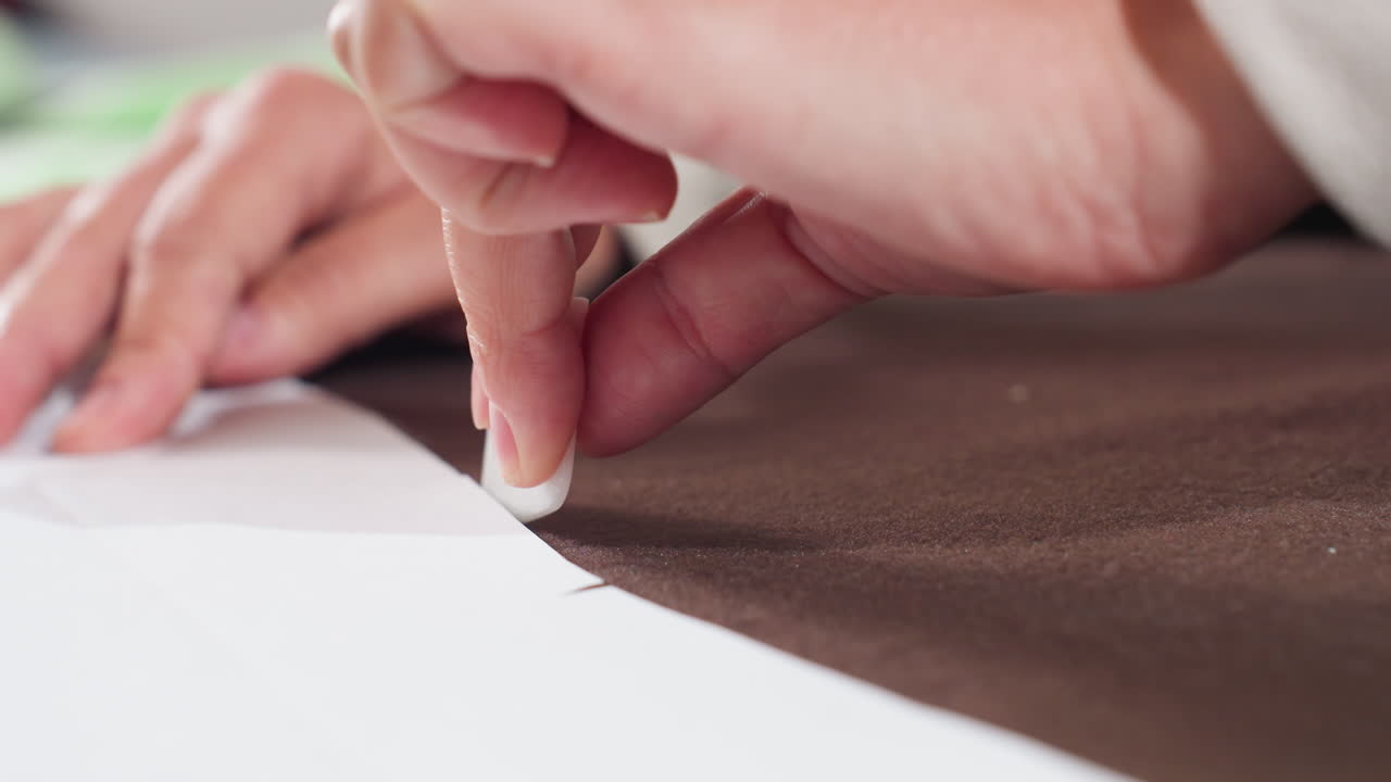 Designer holding cloth firmly while using block shaped tailor chalk to carefully draw pattern outline on thick brown fabric for garment making during creative process inside sewing workshop