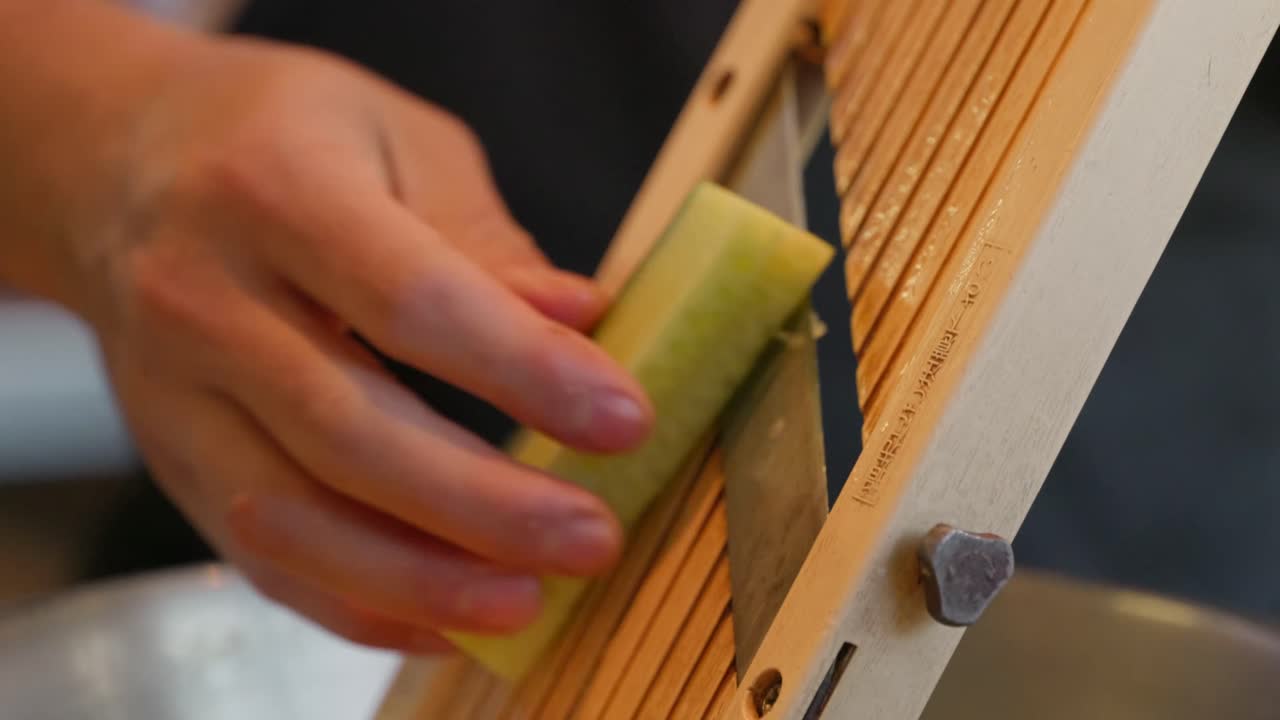 A chef slices a cucumber on a mandolin without using a safety guard, flipping it and turning it as he goes in an up and down motion