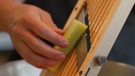 a chef slices a cucumber on a mandolin without using a safety guard, flipping it and turning it as he goes in an up and down motion