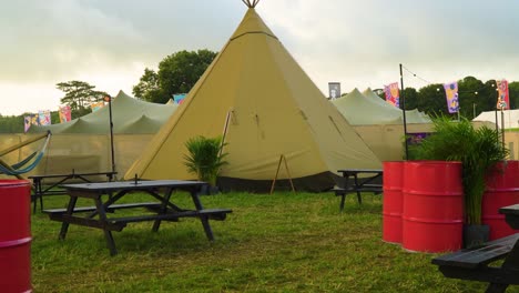 relaxation zone on summer music festival tilting camera movement revealing tipi tent in with red barrels with plants on grassy field high class camping glamping flags in the background sunset