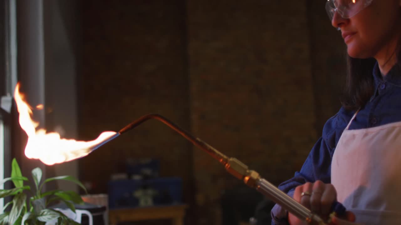 가스 버너 를 사용 하는 아프론 과 안경 을 입고 작업장 에 있는 백인 여성 보석업자