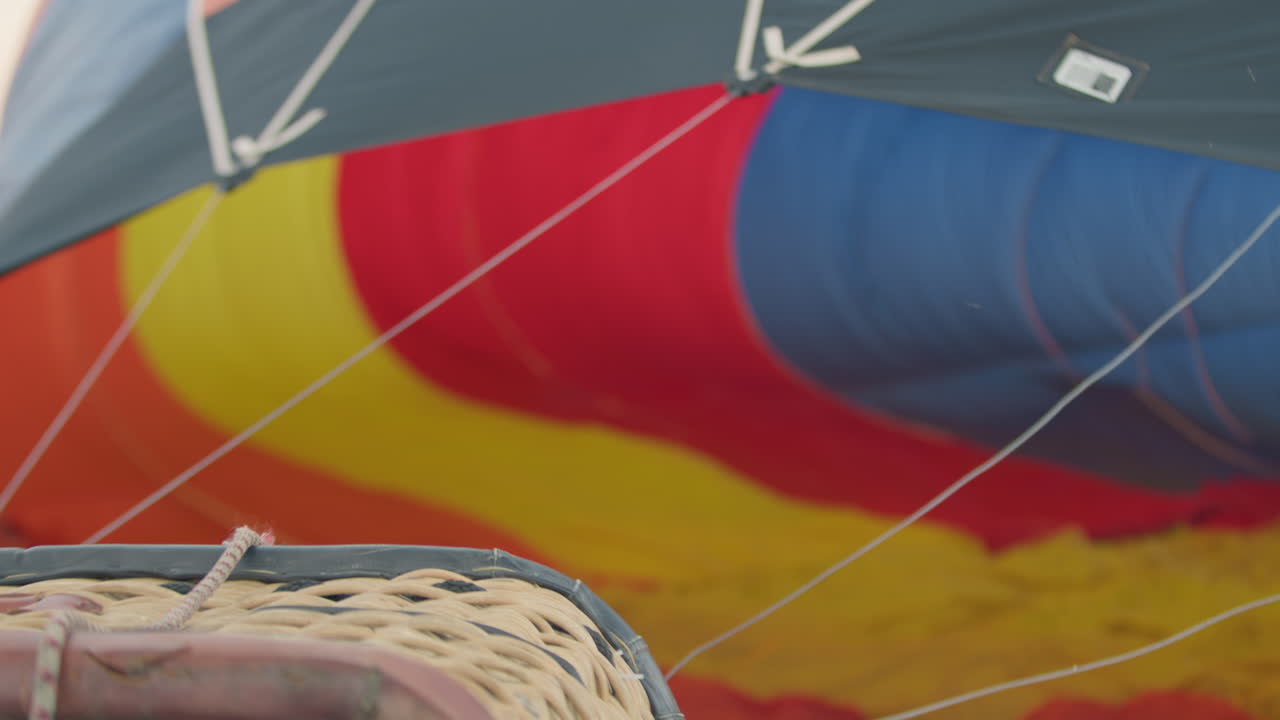 Detailed view of vibrant red, yellow, and blue hot air balloon fabric inflating in background while woven wicker basket with attached ropes remains in foreground during outdoor launch setup