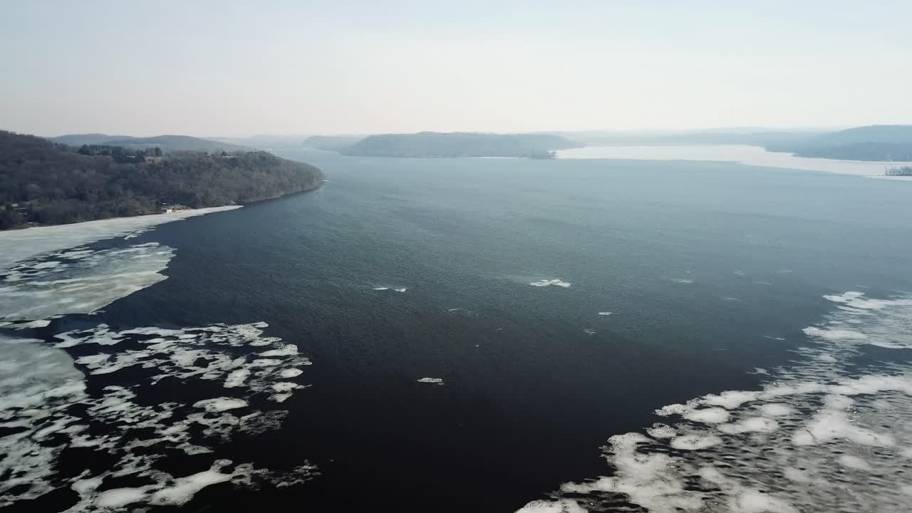 A wide shot of a river or lake with ice floes during winter, surrounded by forested banks and hills under an overcast sky.