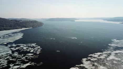 a wide shot of a river or lake with ice floes during winter, surrounded by forested banks and hills under an overcast sky.