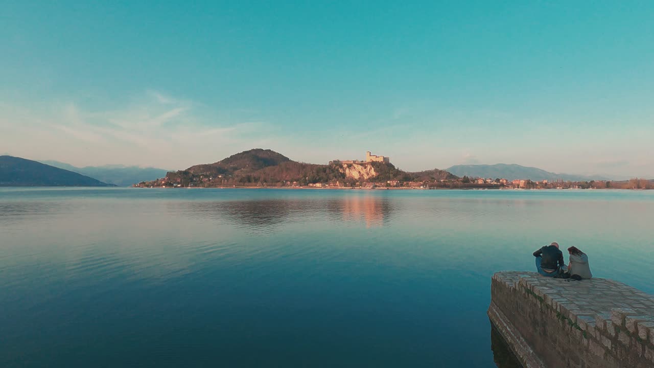 Romantic couple sit on jetty edge over Maggiore lake and look at Angera castle in background at sunset, Italy