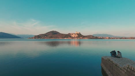Romantisches-Paar-Sitzt-Am-Steg-über-Dem-Lago-Maggiore-Und-Blickt-Bei-Sonnenuntergang-Auf-Das-Schloss-Angera-Im-Hintergrund,-Italien