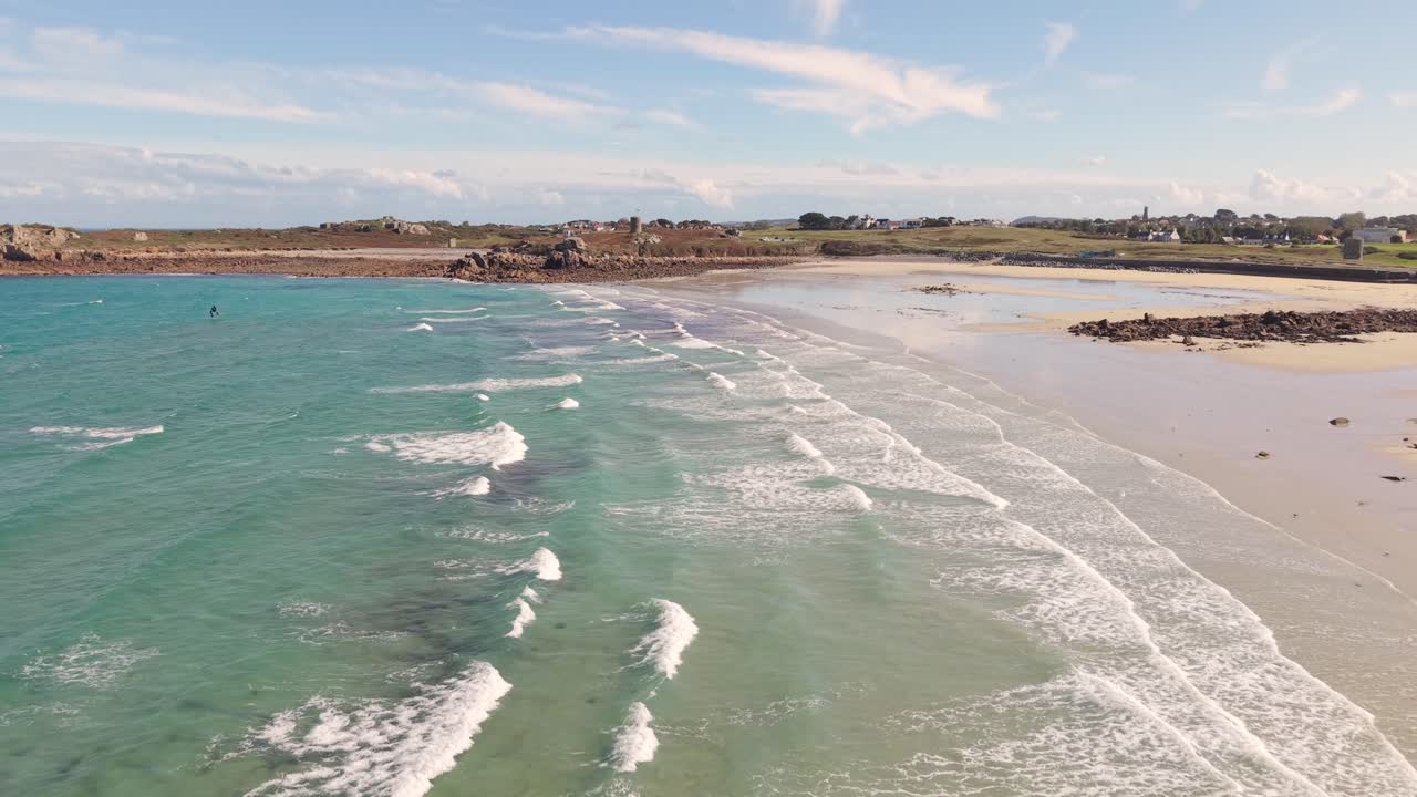 Low flight over waves coming ashore at Lancresse Bay Guernsey with golden sandy beach rocky outcrops crystal clear turquoise sea on sunny day