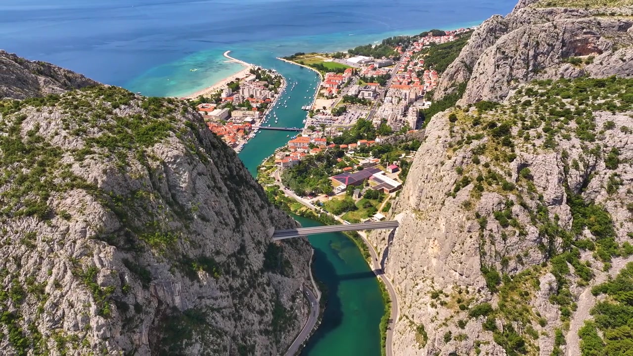 Omiš se nachází tam, kde se řeka vlévá do moře. Řeku překlenuje most. Budovy lemují pobřeží. Na vodě jsou lodě. Pobřeží má pláže a skály.
