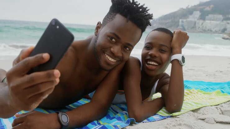 Coppia afroamericana ripresa frontalmente che si scatta un selfie con il cellulare sulla spiaggia 4K