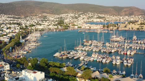 aerial drone flying across the bodrum marina with large sailboats docked in the aegean sea of mugla turkey as the sun sets over the hills and white villas on a summer afternoon