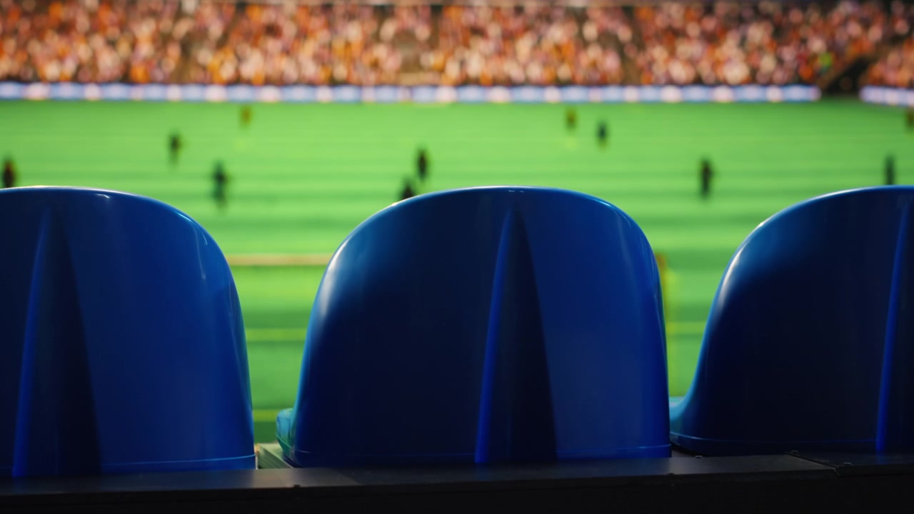 Primer plano de asientos azules de estadio y campo de fútbol borroso con jugadores