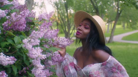 black lady seductively smells touches pink lilac flowers in park, eye contact