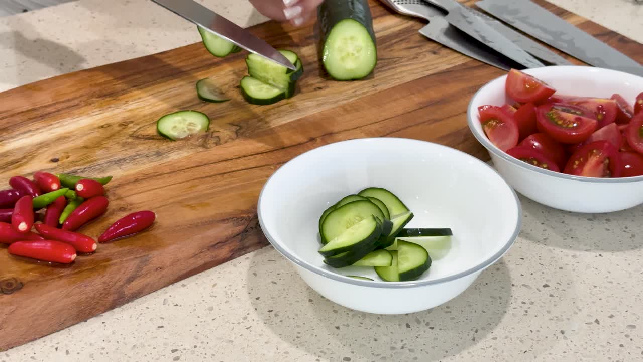A person slices cucumber on a wooden cutting board with tomatoes and red chili peppers, transferring pieces into a white bowl under bright, natural kitchen lighting