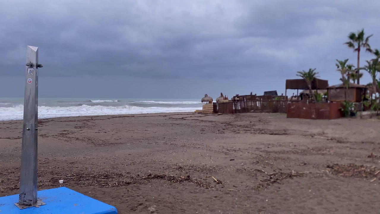 Prázdná plážová sprcha stojící na pláži s tmavým pískem, s rozbouřenými oceánskými vlnami a opuštěným plážovým barem pod zataženou oblohou.